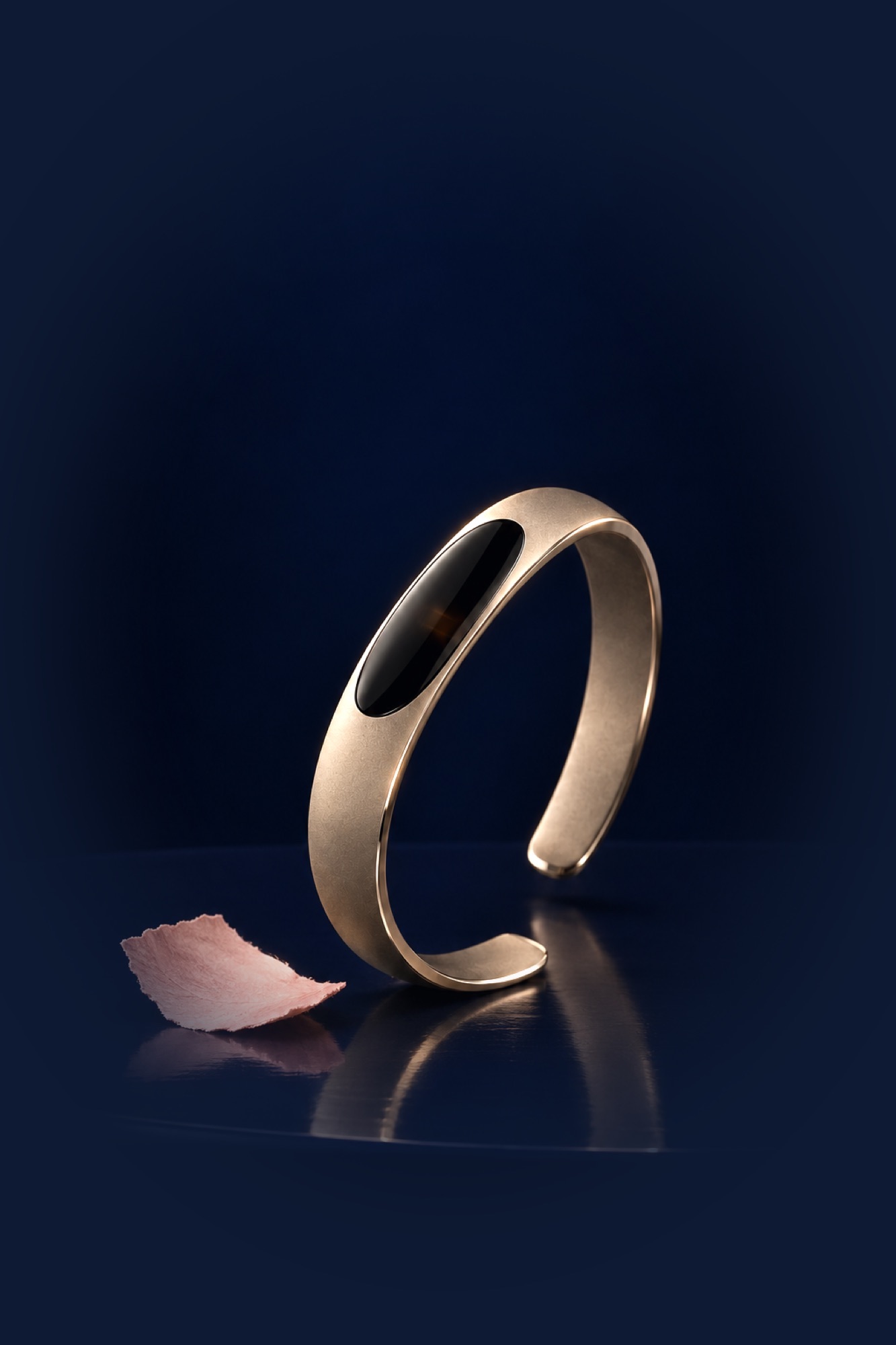 Le Pinson in champagne titanium under a single warm light, a blush paper petal beside it